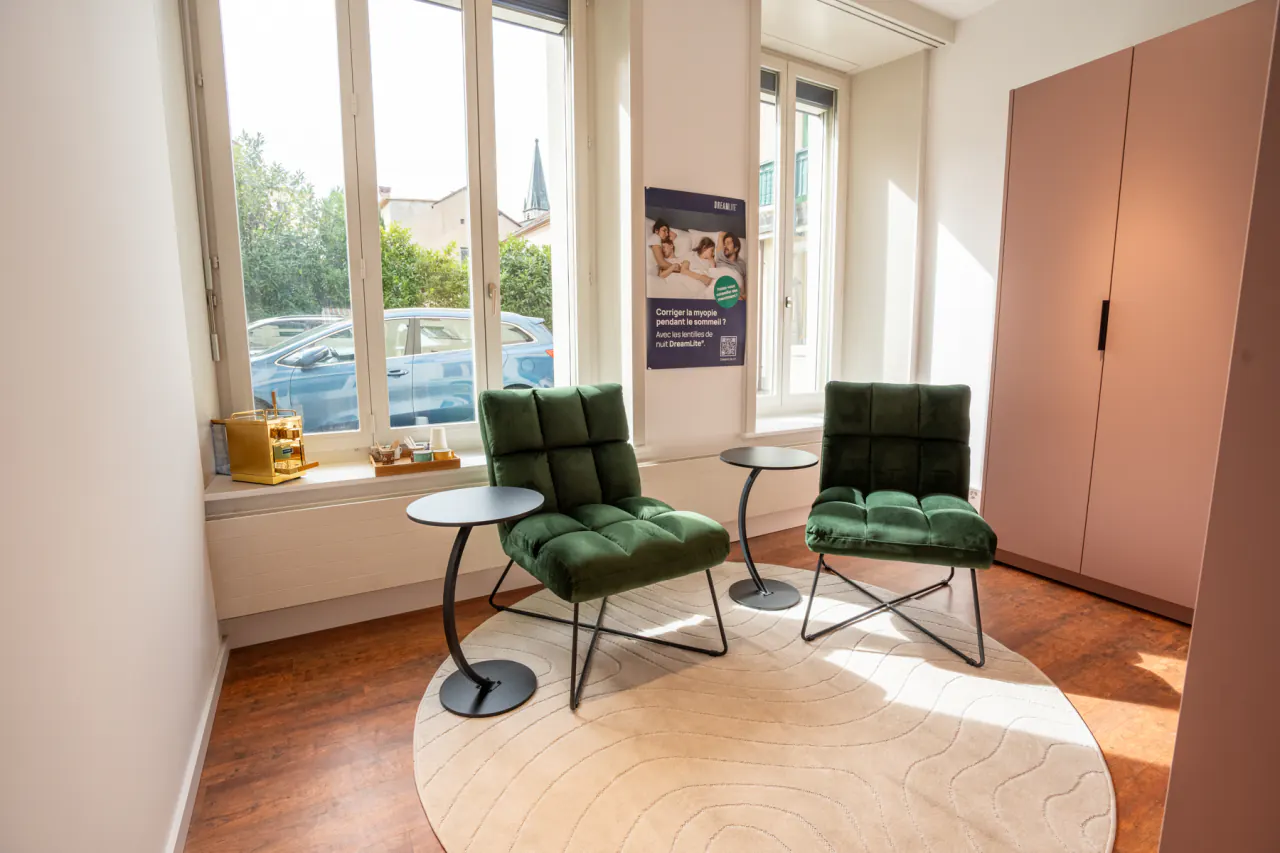 Coin salon du magasin d’optique à Payerne : deux fauteuils en velours vert et leurs guéridons, sur un tapis rond clair, devant les fenêtres