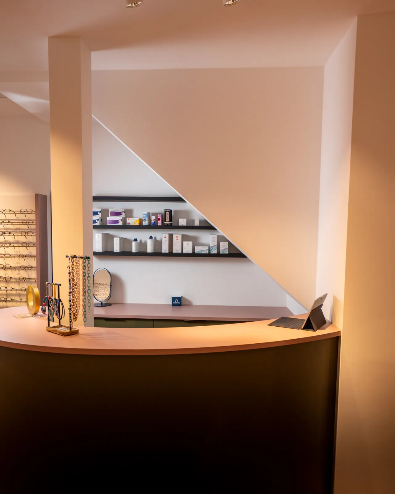 Comptoir d’accueil de l’opticien à Payerne : un plan de travail courbe rose, un présentoir de chaînes à lunettes et les produits d’entretien alignés sur deux étagères