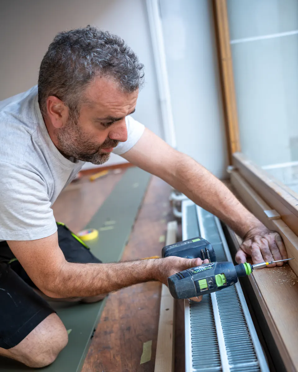 Finitions au pied de la vitrine : un artisan visse à la perceuse, au-dessus d’un radiateur