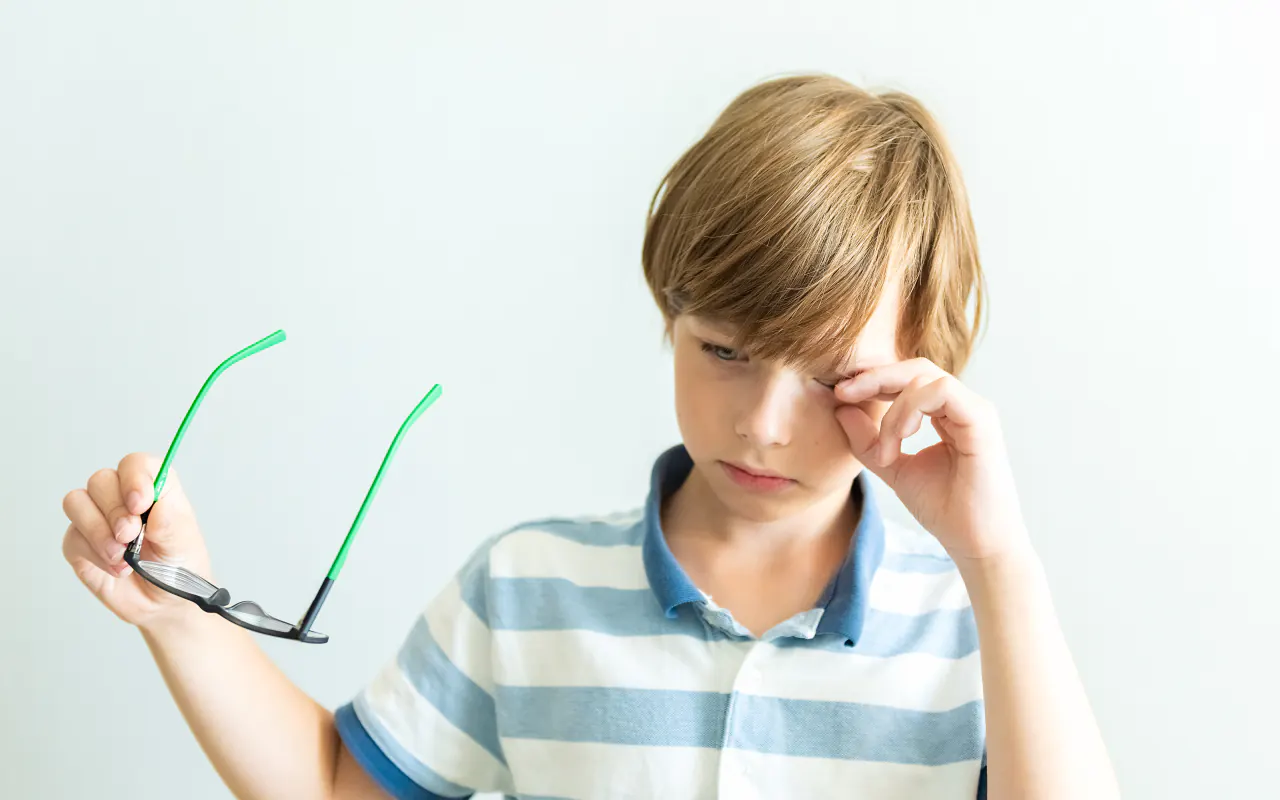 Freination de la myopie chez l’enfant : jeune garçon se frottant l’œil, ses lunettes à la main