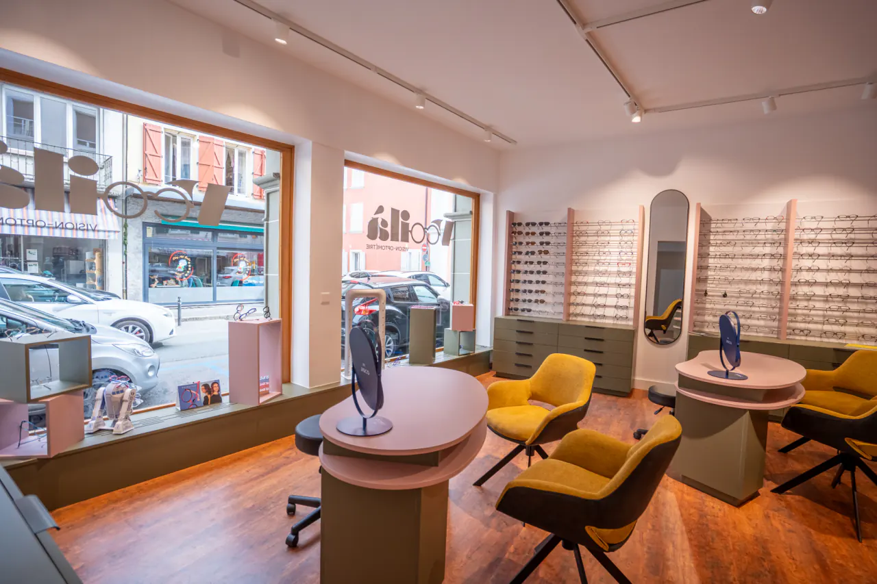 Magasin d’optique à Payerne : les murs de montures au fond, deux tables d’essayage rondes et des fauteuils jaunes, face à la grande vitrine ouverte sur la rue