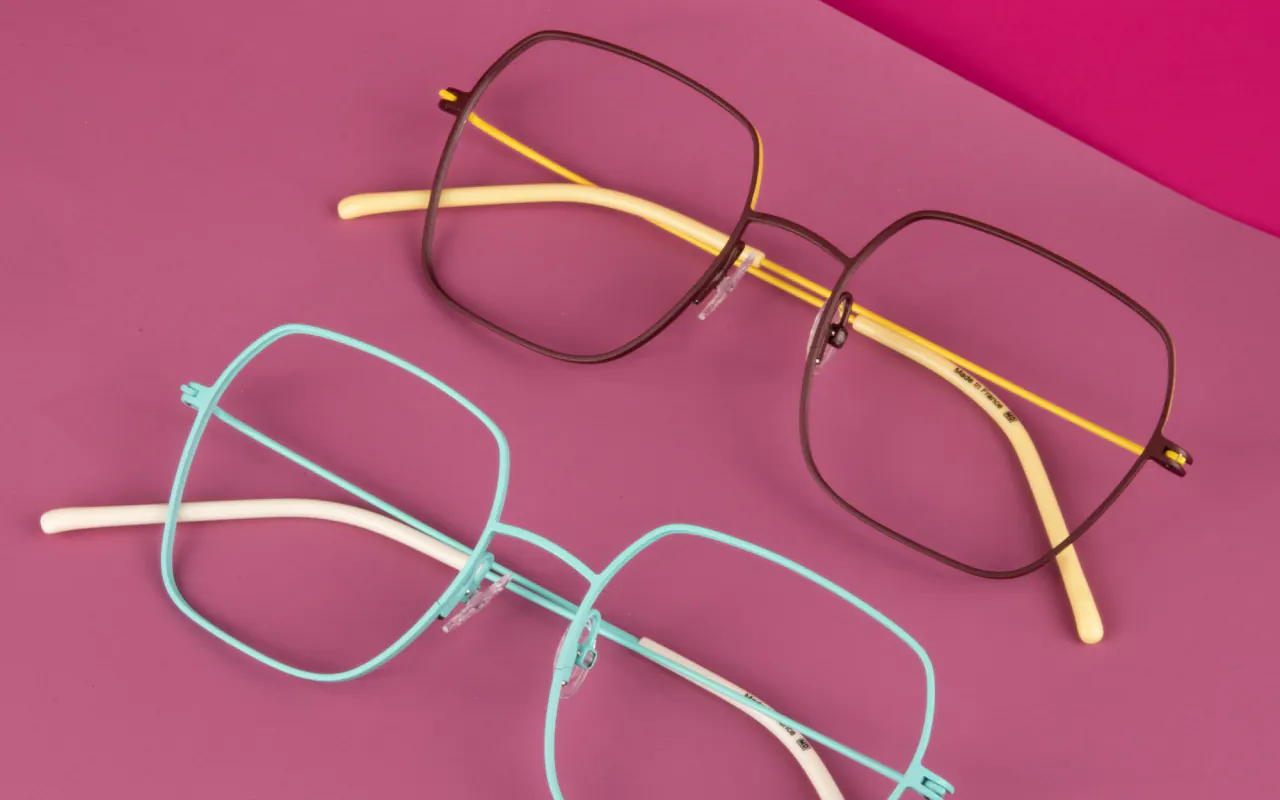 Lunettes et montures : deux montures hexagonales en métal fin, l’une brune à branches jaunes, l’autre turquoise, posées sur un fond rose