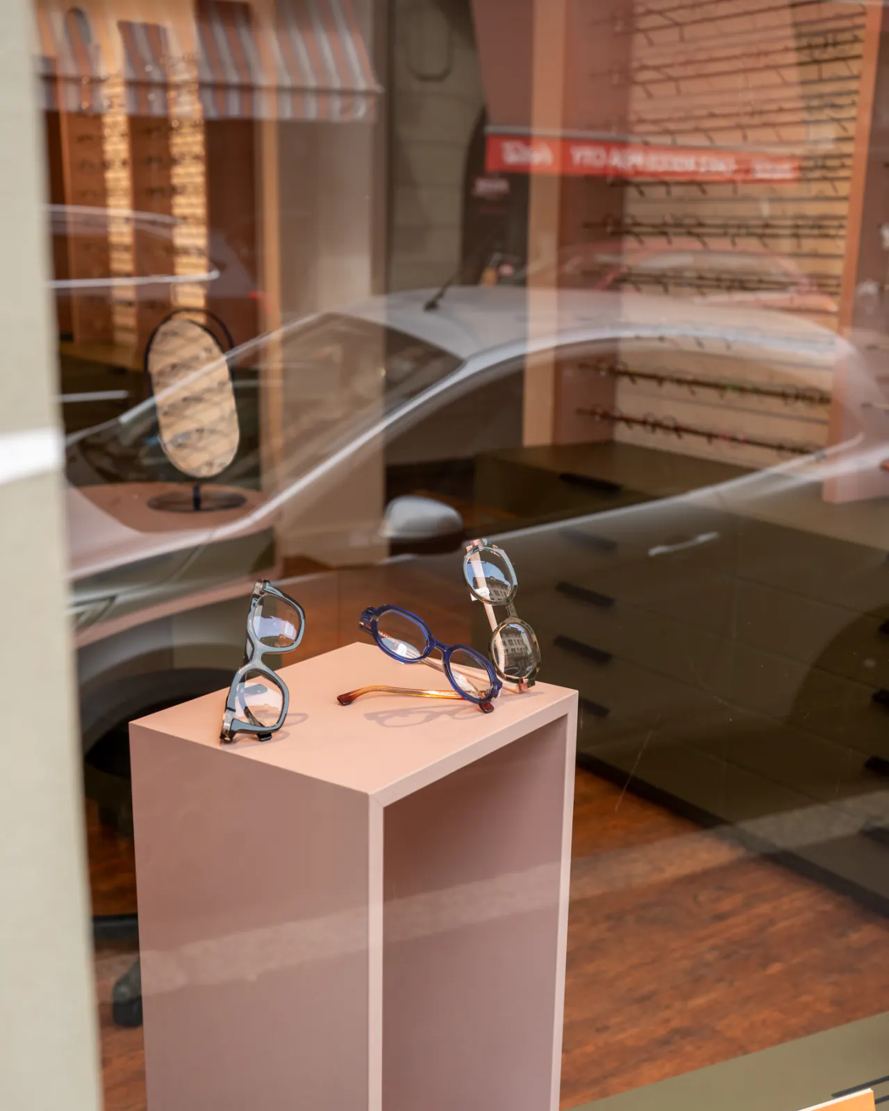 Montures en vitrine : trois paires de lunettes posées sur un socle rose, derrière la glace où se reflète la rue de Lausanne