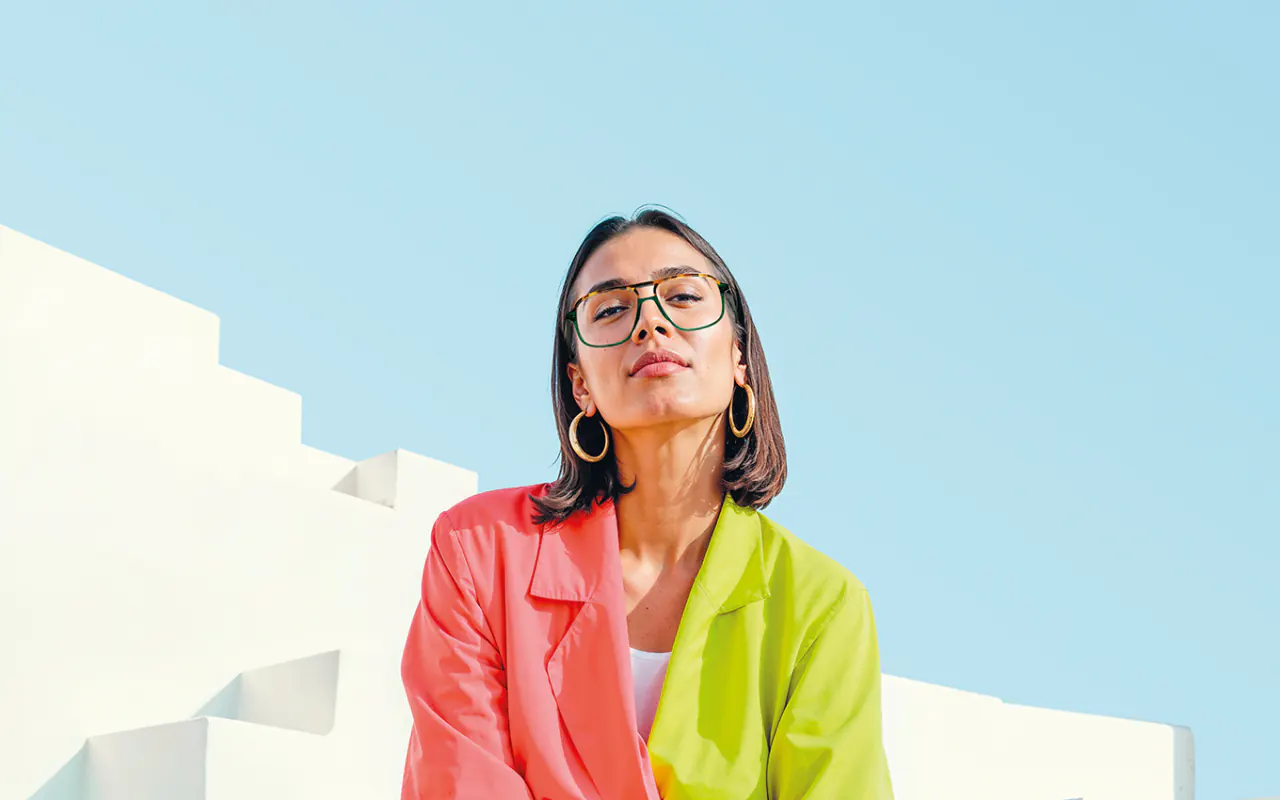 Offres lunettes : monture fine Jisco verte et écaille à double pont, portée avec une veste rose et vert anis devant un ciel bleu et des marches blanches