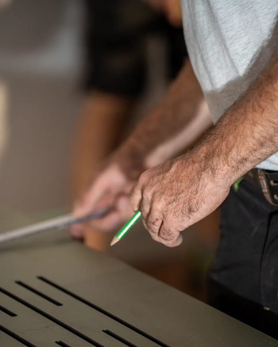 Traçage au crayon vert sur un panneau anthracite ajouré, gros plan sur les mains d’un menuisier