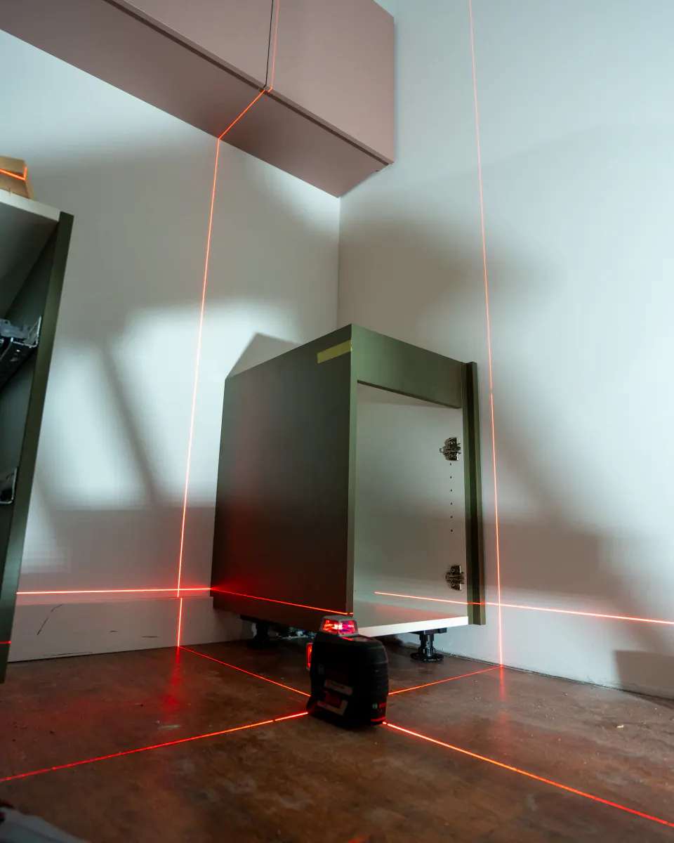 Pose des meubles au laser : lignes rouges sur le sol et les murs, autour d’un caisson vert et d’un meuble rose suspendu