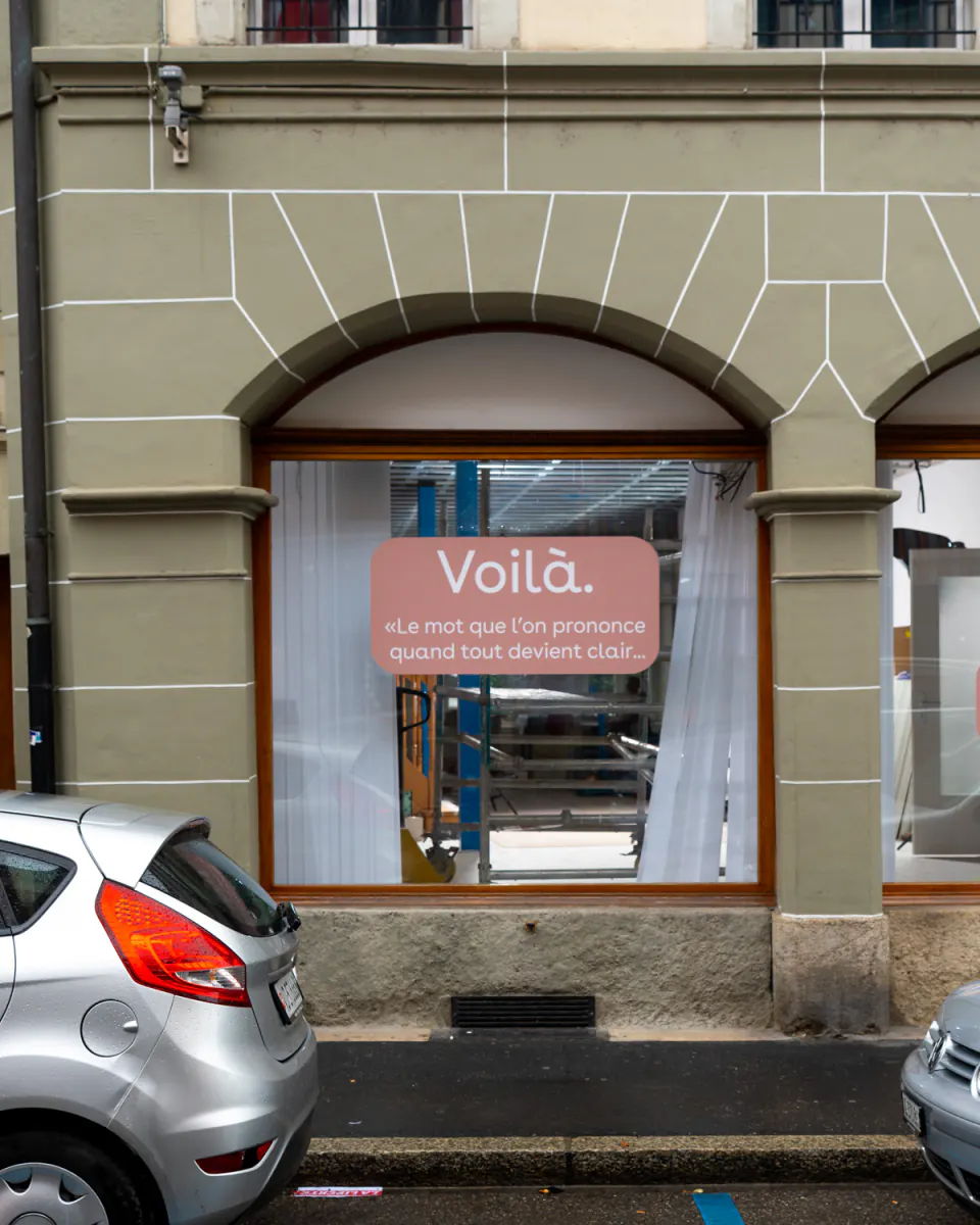 Vitrine du futur magasin d’optique Voilà à Payerne pendant les travaux : sous l’arcade, une vitrophanie rose annonce « Voilà. Le mot que l’on prononce quand tout devient clair… »