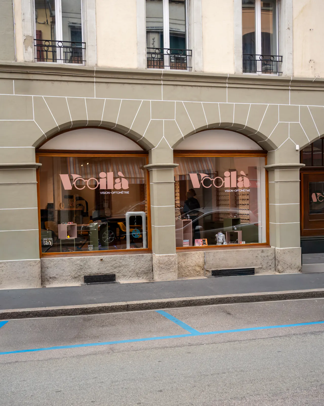 Vitrine de l’opticien Voilà à Payerne : la façade vert sauge du 42 rue de Lausanne, ses deux arcades et le nom « Voilà Vision-Optométrie » en rose sur les vitres